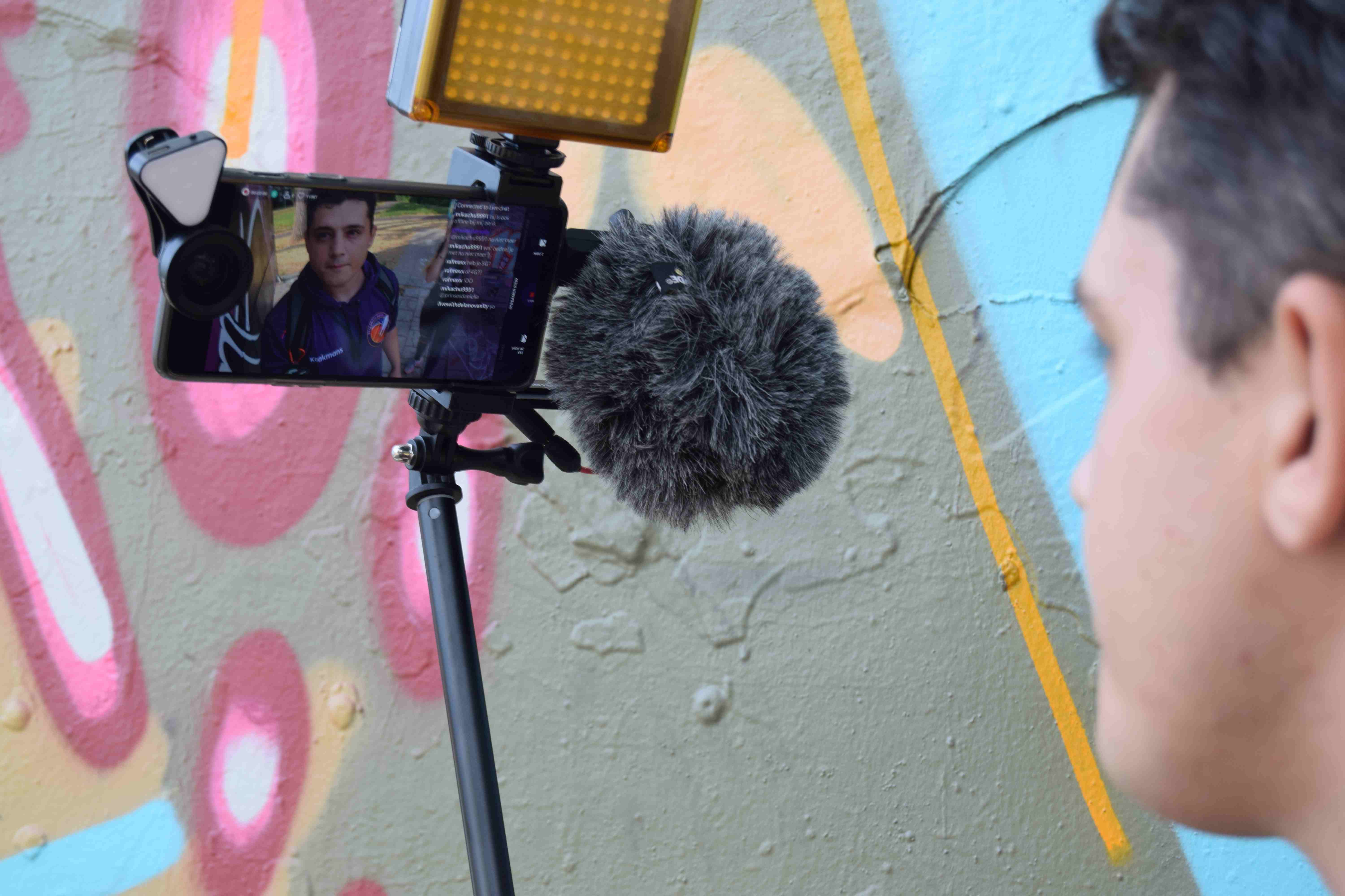 Kaakmans aan het IRL streamen op straat met telefoon, shotgunmicrofoon en LED-licht voor een graffitimuur